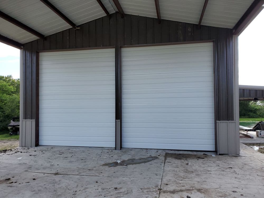 Two large white roll-up garage doors on a metal building with a concrete floor.