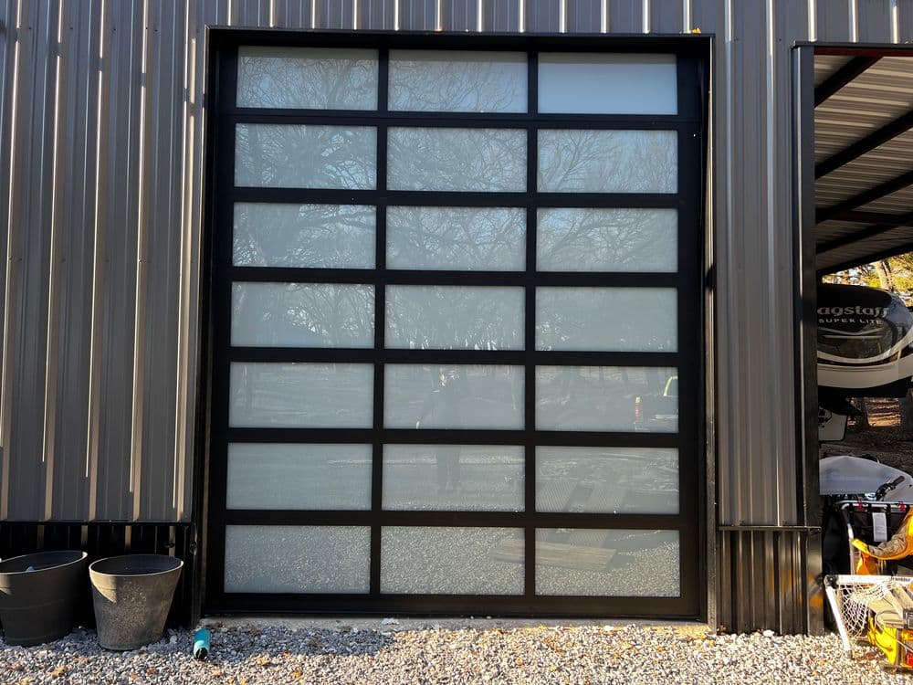 Modern black framed glass garage door on a metal building, surrounded by gravel and pots.