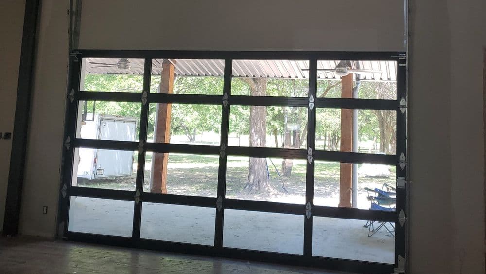 Glass garage door open to a view of trees and outdoor seating area. Bright natural light.