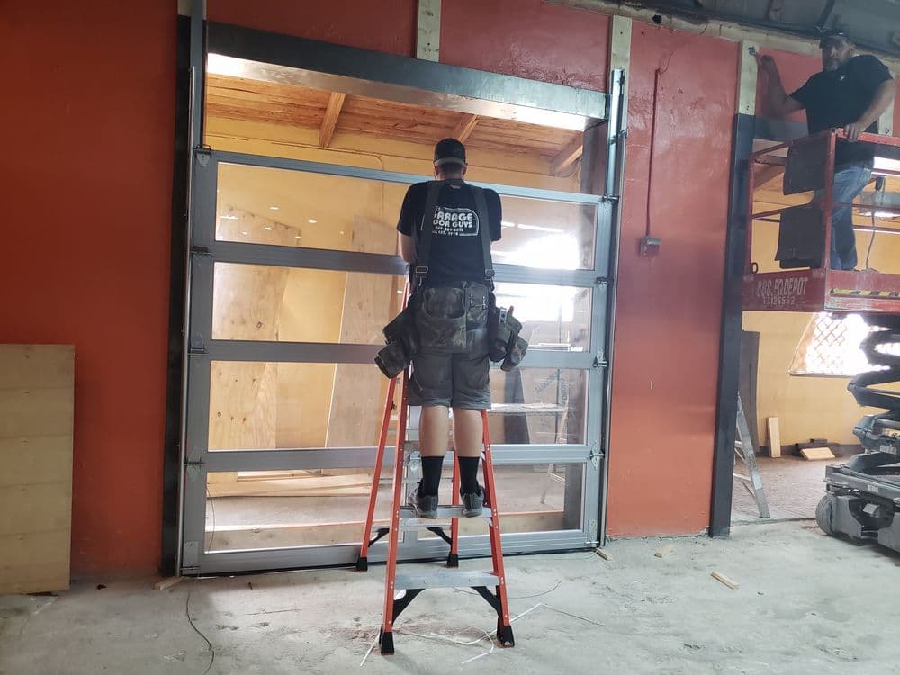 Worker installing a glass garage door while using a ladder in a construction site.