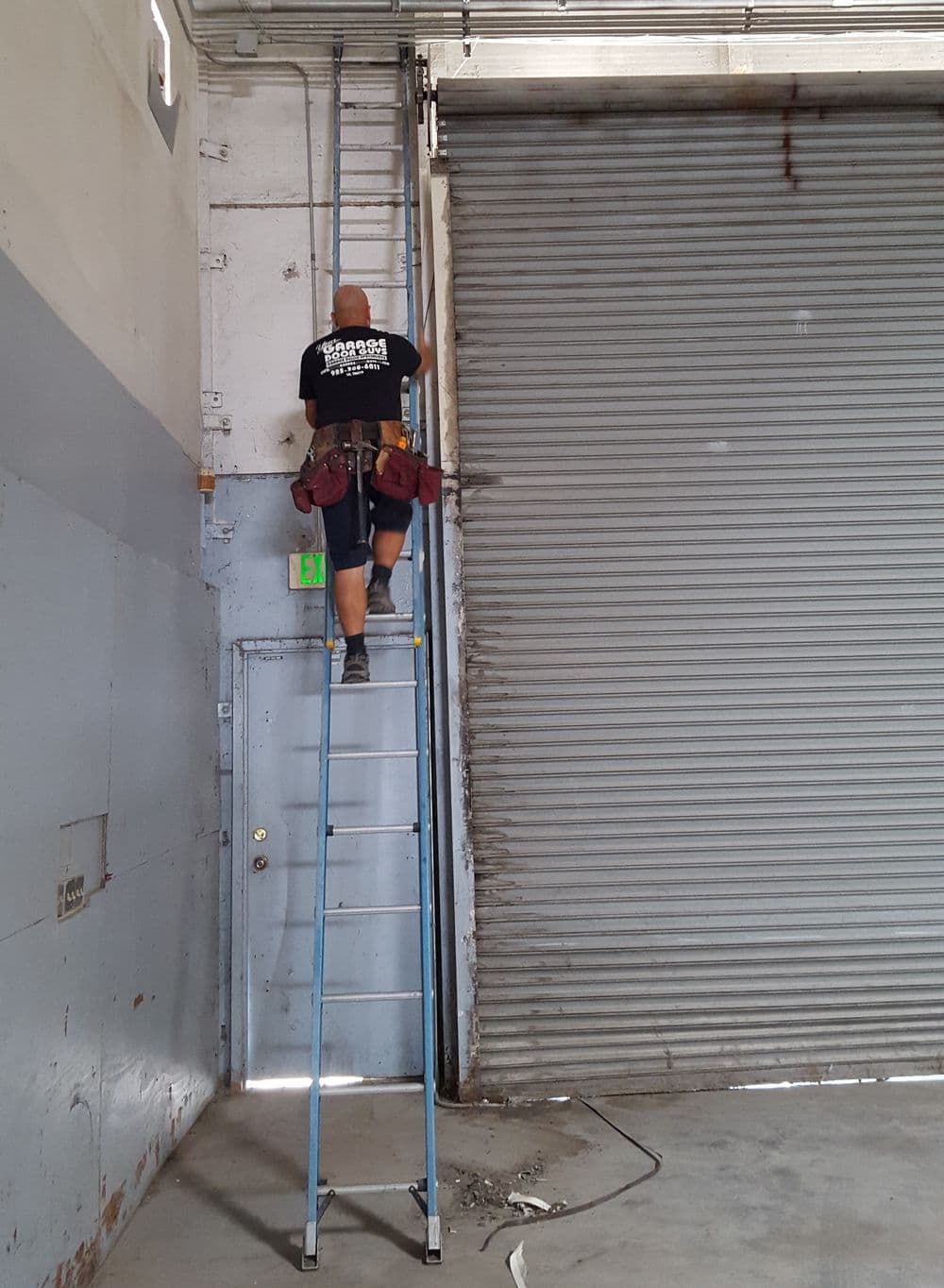 Worker on a ladder performing maintenance in a warehouse with a roll-up door.