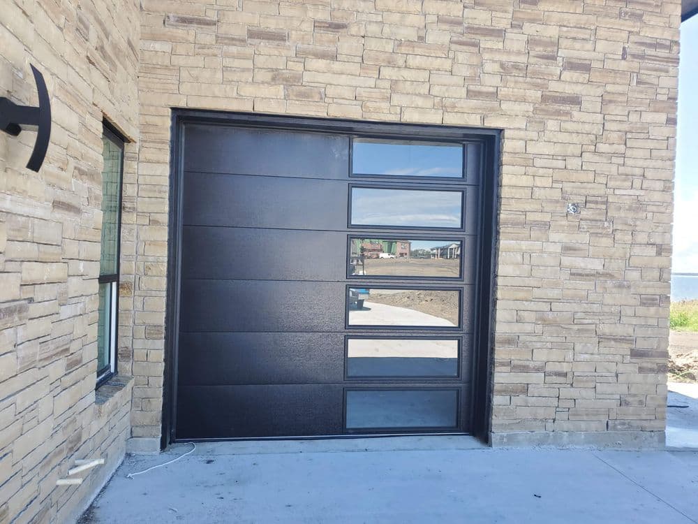 Modern black garage door with large glass panels on a stone exterior home.