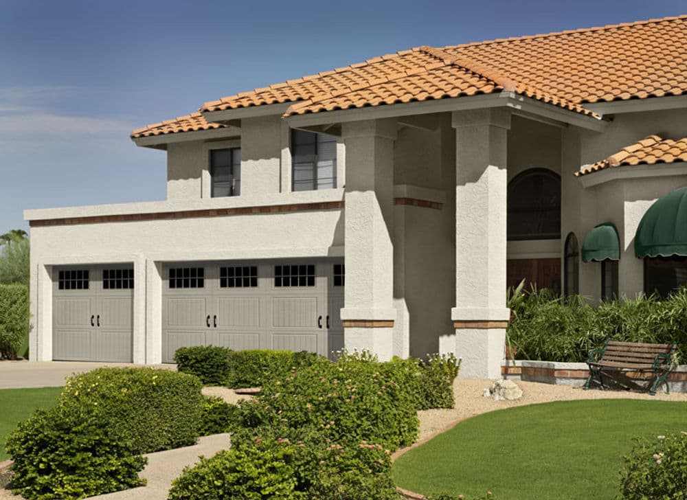 Modern home exterior with beige stucco, tile roof, and stylish garage doors. Lush landscaping.
