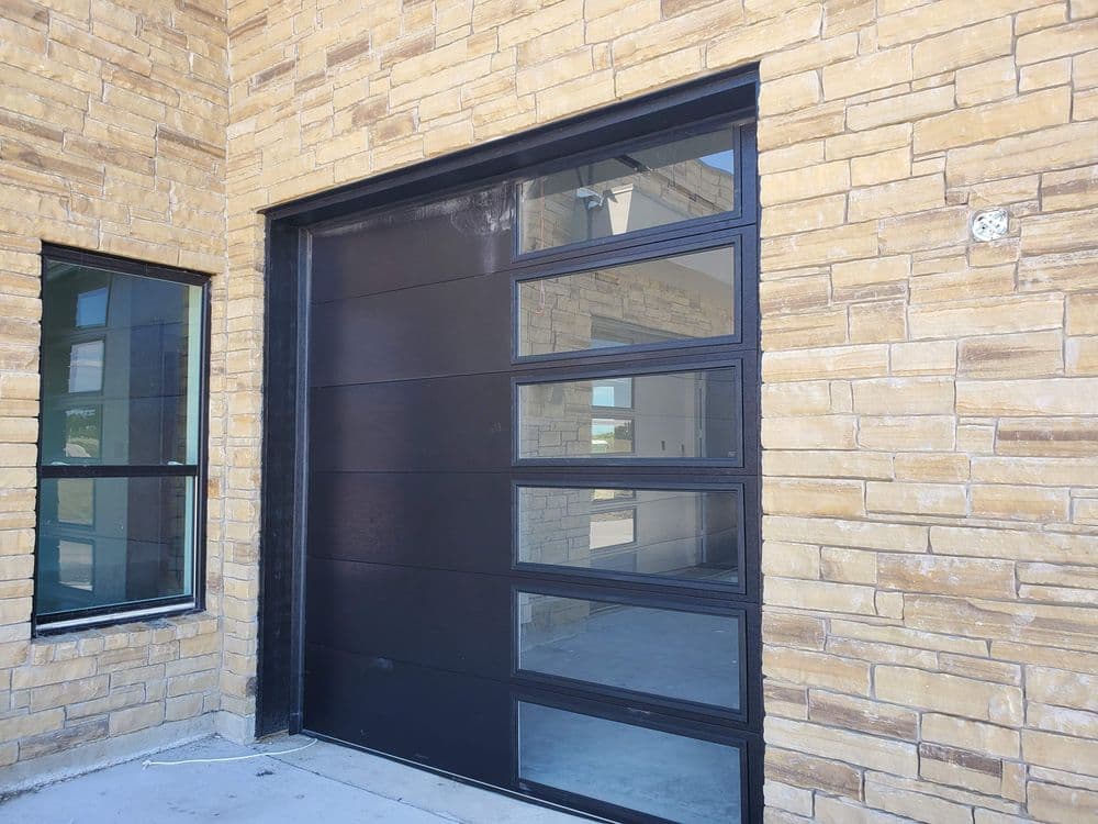 Modern black garage door with large windows, set against a textured stone wall.