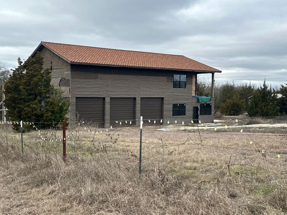Two-story house with brown garage doors, surrounded by dry grass and trees under a cloudy sky.