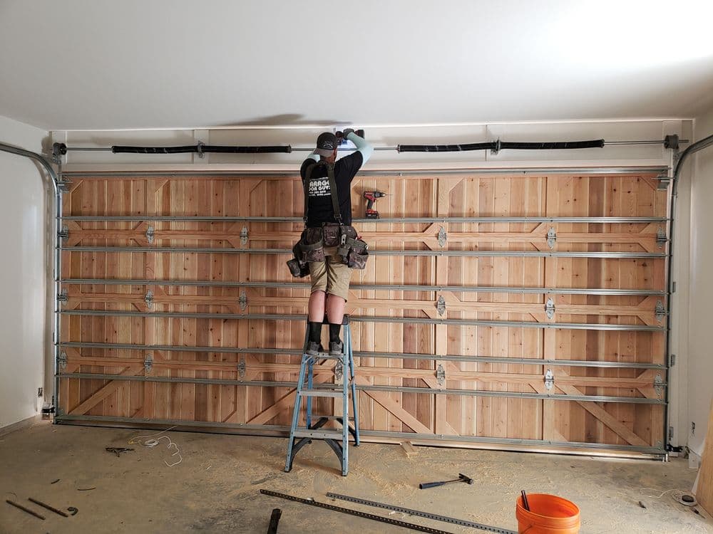 Person installing a wooden garage door on a DIY project using a ladder and power tools.